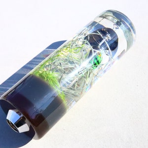 May include: A clear cylindrical glass gear stick knob with a green and black fishing lure inside. The bottom of the knob is black and the top is clear.