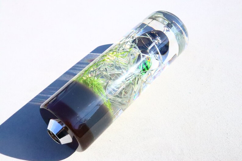 May include: A clear cylindrical glass gear stick knob with a green and black fishing lure inside. The bottom of the knob is black and the top is clear.