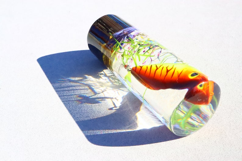 Może przedstawiać: Przezroczysty szklany cylinder ze srebrnym wierzchołkiem, zawierający kolorową szklaną rybę i zielone i fioletowe szklane rośliny. Cylinder leży na boku na białej powierzchni.