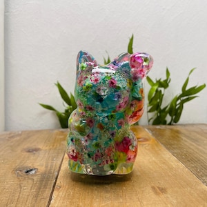 Puede incluir: Una colorida figura de Maneki Neko de resina. El gato está decorado con un estampado floral en tonos rosa, verde y azul. El gato de la suerte está posando con una pata levantada, un gesto tradicional de bienvenida y buena fortuna.