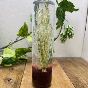 May include: A clear glass cylinder vase filled with dried white flowers and a red base. The vase is sitting on a wooden surface.