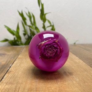 May include: A magenta sphere with a preserved rose at its centre. The rose is a deep burgundy colour, encased within the clear, glossy orb. The sphere sits on a wooden surface, with green foliage in the background.