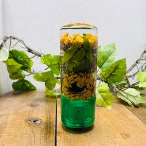 May include: A clear glass cylinder filled with dried flowers and leaves, with a green base. The cylinder is sitting on a wooden surface.