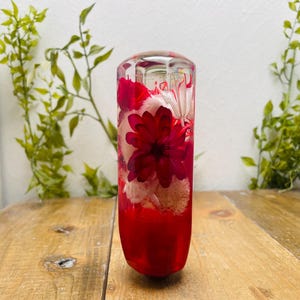 Könnte beinhalten: Ein klarer, zylindrischer Gegenstand mit roter Basis, gefüllt mit roten und weißen Blumen. Die Blumen sind in einem transparenten Material eingeschlossen, wodurch ein dekoratives Stück entsteht. Der Gegenstand steht auf einer Holzoberfläche.