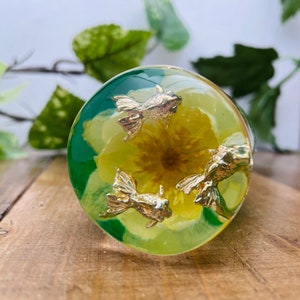以下が含まれることがあります： 黄色い花と金色の魚が3匹入った、丸い透明なレジン製のペーパーウェイト。ペーパーウェイトは緑色の背景です。