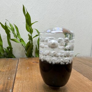 Puede incluir: Un jarrón de vidrio transparente con una base marrón oscuro, lleno de burbujas transparentes y cuentas blancas con forma de perla. Ramas verdes y frondosas son visibles en el fondo. El jarrón está sobre una superficie de madera.