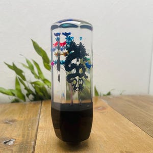 May include: A clear, cylindrical gear stick knob featuring a black dragon design and colourful floral details. The base is black, and the top has a faceted design. The knob is displayed on a wooden surface.