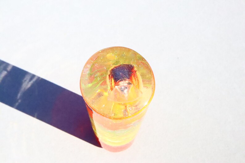 May include: A clear cylinder filled with layers of colorful glitter and resin. The top layer is yellow with green glitter, the middle layer is orange, and the bottom layer is red. A small, dark brown object is embedded in the resin.