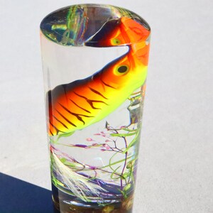 Może przedstawiać: Przezroczysty szklany cylinder z kolorową przynętą wędkarską w środku. Przynęta jest pomarańczowa, ż&oacute;łta i czarna z białym paskiem. Cylinder jest wypełniony przezroczystą cieczą i ma niewielką ilość zielonego i fioletowego materiału roślinnego na dnie.