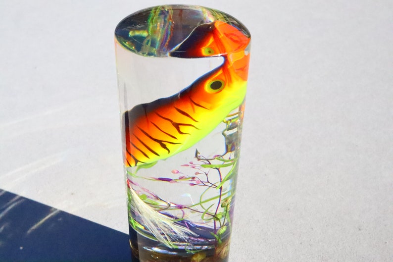 Może przedstawiać: Przezroczysty szklany cylinder z kolorową przynętą wędkarską w środku. Przynęta jest pomarańczowa, ż&oacute;łta i czarna z białym paskiem. Cylinder jest wypełniony przezroczystą cieczą i ma niewielką ilość zielonego i fioletowego materiału roślinnego na dnie.