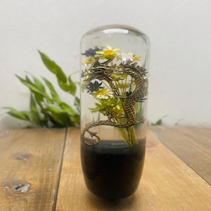 May include: A clear glass decorative piece with a bronze dragon wrapped around a bouquet of white and yellow flowers. The bottom of the glass is filled with a dark substance. The piece is cylindrical and sits on a wooden surface.