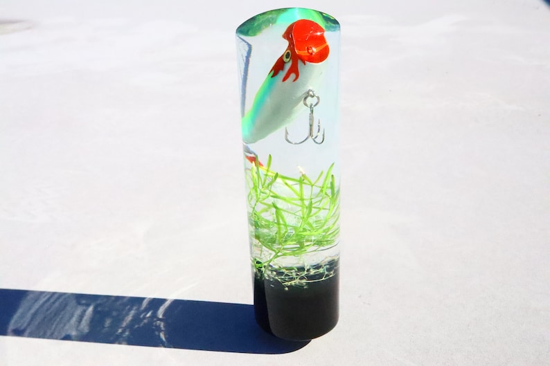 Pu&ograve; includere: Un cilindro trasparente riempito di resina, contenente un'esca da pesca verde, bianca e rossa con un amo. Anche materiale vegetale verde simile a piante &egrave; incorporato nella resina.