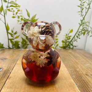 Könnte beinhalten: Eine transparente Bärenfigur aus Kunstharz, gefüllt mit getrockneten Blumen, Tannenzapfen und anderen natürlichen Elementen. Der Bär hat einen rotbraunen Sockel und klare Ohren. Die Figur steht auf einer Holzoberfläche mit Grünzeug im Hintergrund.
