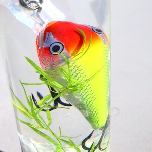 May include: A yellow, red and white fishing lure with a single hook is submerged in a glass of water with green plastic plants.
