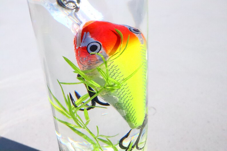 May include: A yellow, red and white fishing lure with a single hook is submerged in a glass of water with green plastic plants.