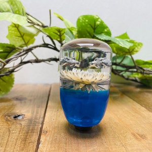 May include: A clear glass gear shift knob with a white flower and two silver fish inside. The bottom of the knob is filled with blue liquid.