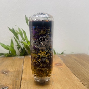 May include: A clear, rectangular bottle with a decorative interior. The bottle contains gold leaf, purple and black accents, and a silver-colored crown design. The bottle is displayed on a wooden surface with green foliage in the background.