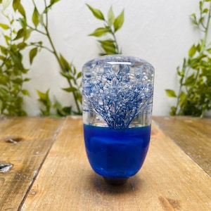 Könnte beinhalten: Ein klarer Schaltknauf aus Harz mit blauen Schleierkrautblüten und weißen Akzenten. Die untere Hälfte ist ein kräftiges, leuchtendes Blau. Der Knauf steht auf einer Holzoberfläche, mit grünem Laub im Hintergrund.