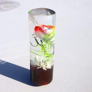 May include: A clear cylindrical tube filled with resin, containing a red, white and green fishing lure, green plastic plants, and a white flower.