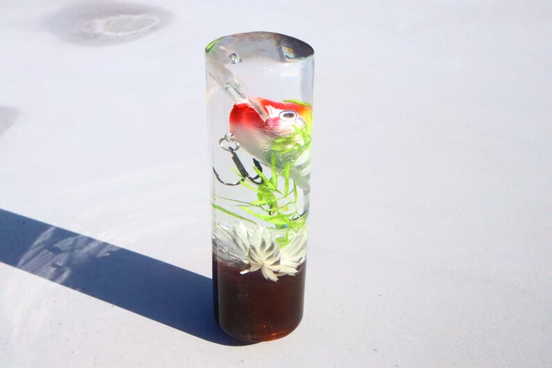 May include: A clear cylindrical tube filled with resin, containing a red, white and green fishing lure, green plastic plants, and a white flower.