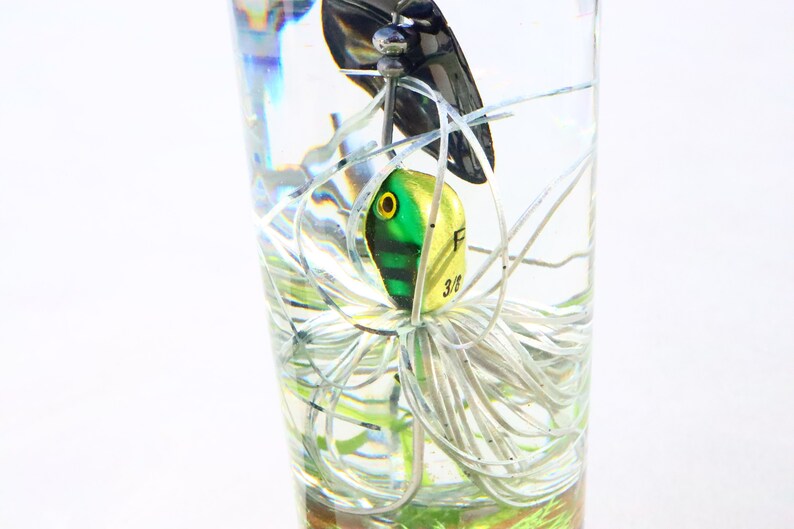 May include: A green and yellow fishing lure with white and silver fishing line in a clear glass. The lure has a black eye and the text "F 3/8" on the side.