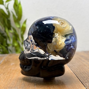 May include: A black skull-shaped decorative object with a clear top filled with dried yellow and blue flowers. The skull is sitting on a wooden surface.