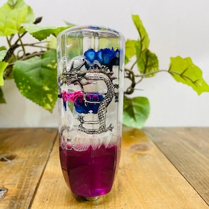 Könnte beinhalten: Ein transparenter Glas-Schalthebelknauf mit einem silbernen Drachen und rosa und blauen Blumen im Inneren. Die Basis des Knaufs ist mit einem dunkelvioletten Harz gefüllt.