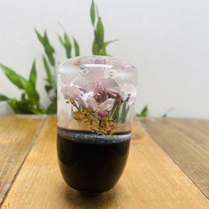 May include: A decorative resin shift knob with a dark base and a clear top. The clear top encases dried pink flowers and small seashells. The shift knob is on a wooden surface with green foliage in the background.