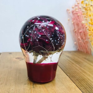 Op de afbeelding: Een heldere glazen bol met een donkerrode bloem en witte accenten erin. De bol is gemonteerd op een donkerrode basis.