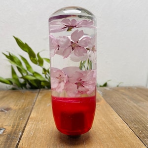Puede incluir: Pomo de palanca de cambios cilíndrico transparente con flores de cerezo rosas encapsuladas en resina. La base es de color rojo vivo. Diseñado para el cambio de marchas de un vehículo. Aproximadamente 10 cm de altura.