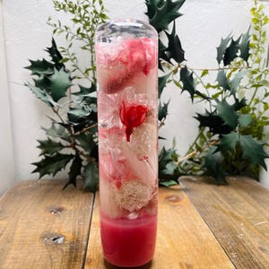 Könnte beinhalten: Ein durchsichtiger Glaszylinder, gefüllt mit rosa und weißen Blumen, die in Harz konserviert sind. Die Blumen sind vertikal im Zylinder angeordnet, mit den rosa Blumen unten und den weißen Blumen oben.