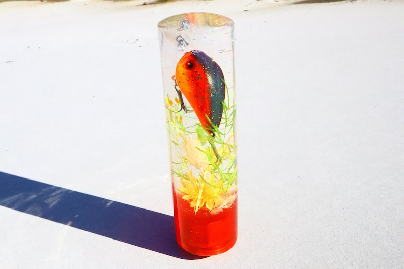 May include: A clear cylinder filled with red liquid and a small orange and black fishing lure. The lure is suspended in the liquid with green and yellow flowers and plants.