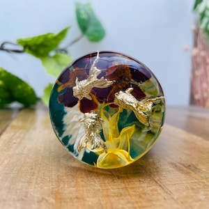 Op de afbeelding: Een ronde, transparante hars-papiersteen met een kleurrijk bloemenontwerp en goudkleurige vissen. De bloemen zijn in tinten paars, geel en wit.