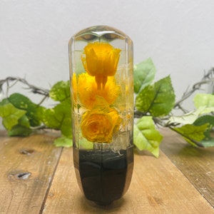 Puede incluir: Una perilla de cambio de marchas de resina transparente con rosas amarillas preservadas en su interior. La perilla tiene una base negra.