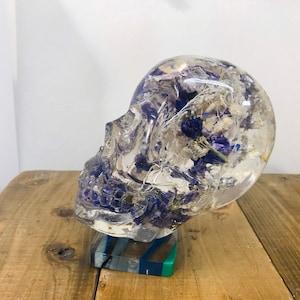 May include: A clear resin skull sculpture with embedded dried flowers in shades of purple and white. The skull is sitting on a multi-coloured base.