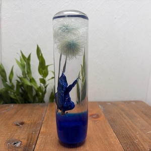 Puede incluir: Un pisapapeles cilíndrico de resina transparente que contiene una cabeza de semilla de diente de león blanca y un elemento en forma de hoja azul oscuro brillante, con una base azul profundo sólida que se funde con la parte superior.