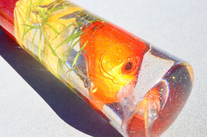 May include: A clear glass cylinder filled with glitter and a red and yellow fish with green plants.