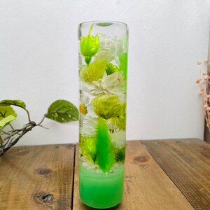 May include: A clear glass cylinder vase filled with preserved green and white flowers. The vase has a green base.