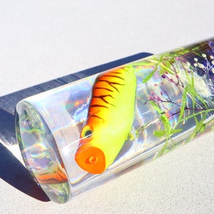 Może przedstawiać: Przezroczysty szklany cylinder wypełniony wodą, ż&oacute;łto-pomarańczową przynętą wędkarską i zielonymi i fioletowymi roślinami. Cylinder znajduje się na białej powierzchni.