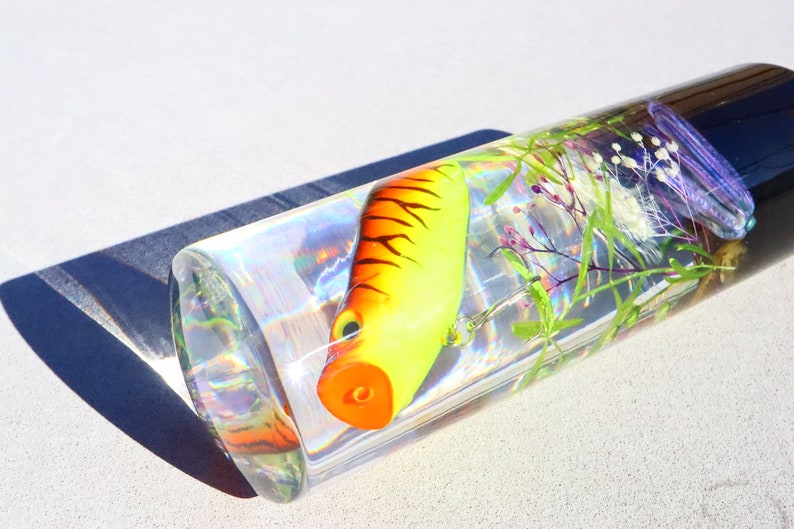 Może przedstawiać: Przezroczysty szklany cylinder wypełniony wodą, ż&oacute;łto-pomarańczową przynętą wędkarską i zielonymi i fioletowymi roślinami. Cylinder znajduje się na białej powierzchni.