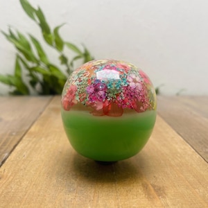 May include: A decorative, round object with a vibrant floral design on top and a bright green base. The floral pattern features pink, red, and teal accents. The object is set on a wooden surface.