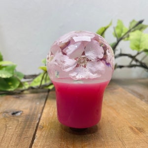 Op de afbeelding: Een roze en transparante resin versnellingspookknop met een ontwerp van witte bloemen die erin zijn geperst.