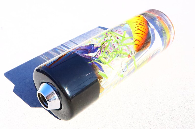 Może przedstawiać: Przezroczysty szklany cylinder z czarną podstawą i chromowanym końc&oacute;wką. Wewnątrz cylindra znajduje się ż&oacute;łto-pomarańczowa przynęta wędkarska, zielone wodorosty i fioletowo-niebieska meduza.