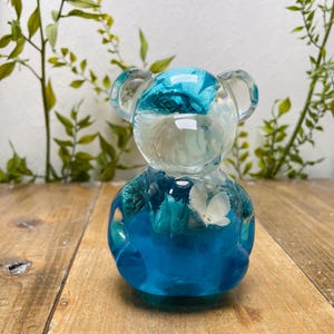 Könnte beinhalten: Eine transparente Bärenfigur aus Kunstharz mit blauen und weißen floralen Einschlüssen. Der Bär hat einen abgerundeten Körper, kleine Ohren und eine glatte, glänzende Oberfläche. Die floralen Elemente sind im Harz sichtbar und erzeugen einen dekorativen Effekt. Die Figur steht auf einer Holzoberfläche.
