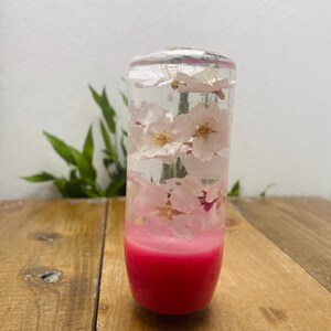 Puede incluir: Un objeto cilíndrico transparente con una base rosa vibrante, lleno de delicadas flores blancas y rosas. Las flores están suspendidas en un líquido transparente, creando una exhibición visualmente atractiva. El objeto está sobre una superficie de madera.