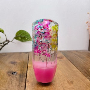 Könnte beinhalten: Ein pinkfarbener und durchsichtiger Schaltknauf aus Harz mit gepressten Blumen im Inneren. Die Blumen sind pink, blau und gelb.