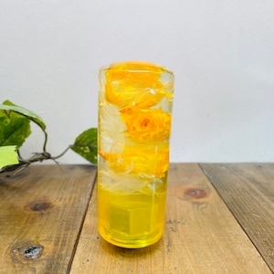 May include: A clear glass cylinder filled with yellow flowers, resembling roses, preserved in resin. The cylinder is on a wooden surface.