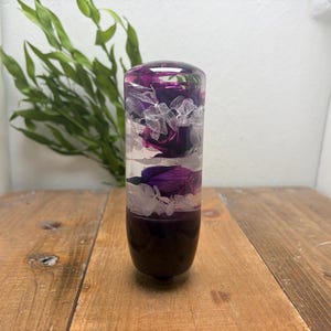 May include: A cylindrical gear shift knob made of clear resin, featuring embedded deep purple flower buds and delicate white dried petals suspended within the material, with a solid dark purple base and a smooth, rounded top surface.
