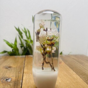 May include: A clear, rectangular resin object with a white base, containing delicate white and pink flowers and brown stems. The floral arrangement is centered within the transparent resin, creating a unique decorative piece. The background features green foliage and a wooden surface.