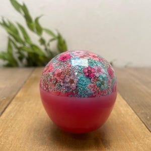 Op de afbeelding: Een bolvormig decoratief object met een bloemenontwerp. De bovenste helft heeft een kleurrijk patroon van roze, blauwe en witte bloemen, terwijl de onderste helft effen helderroze is. Het ligt op een houten oppervlak.